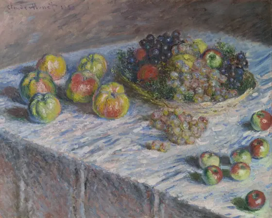 Apples and grapes rest in a shallow wicker basket on a table covered with a light-colored cloth. Additional fruit is scattered on the table.