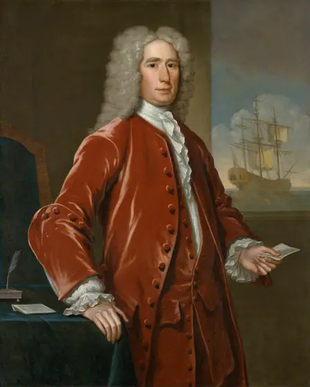 Portrait of a light-skinned man in a long white wig and red-orange coat, holding a folded piece of paper and standing next to a desk, a large sailing ship off his left shoulder.