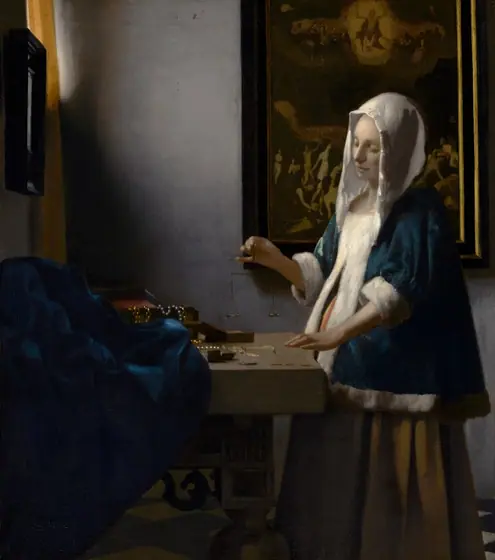 In this vertical painting, a woman stands near the corner of a dimly lit room, facing our left in profile and looking down at a balance she holds suspended in her right hand over a wooden table.