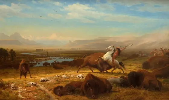 A band of indigenous Americans ride horses toward and through a herd of buffalo, which spreads along a river that winds through plains to mountains in the deep distance in this horizontal landscape painting.
