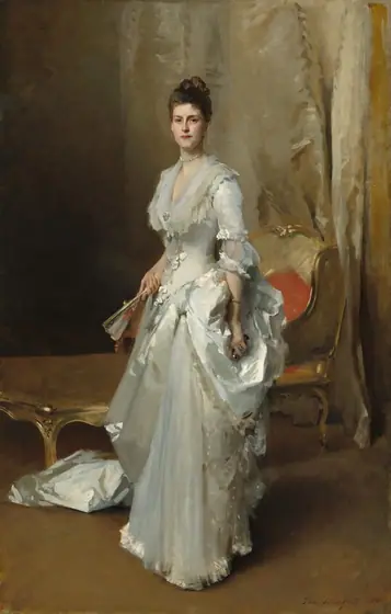 A woman with pale, peachy skin and dark brown hair, wearing a long sleeved, floor-length white satin dress, stands facing and looking out at us in a room hung with shimmering, ivory-white drapery in this vertical portrait.