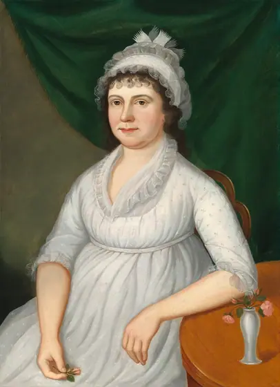 The image is a portrait showing the upper body of a woman seated next to a wooden table. She is positioned facing slightly to the right, with her arm resting on the table and holding a small pink rose in her left hand.
