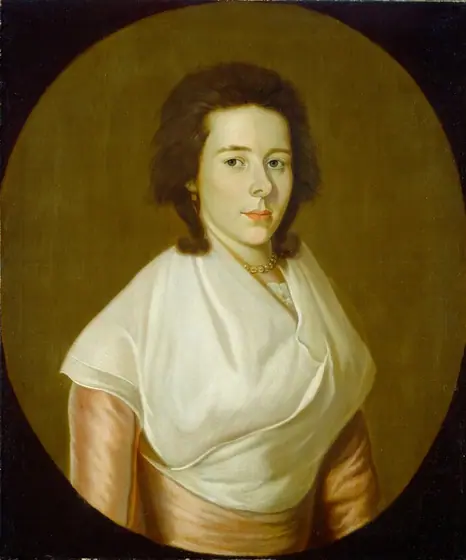 The image shows a woman's upper body. She is looking directly at the viewer with a calm expression. The woman has fair skin, dark brown hair styled away from her face, blue eyes, and delicate features.