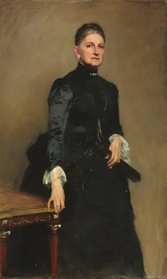 Shown from the shins up, a woman with pale skin, iron-gray hair, and wearing a high-collared, long-sleeved black dress, stands next to a table against a caramel-brown wall in this vertical portrait painting.