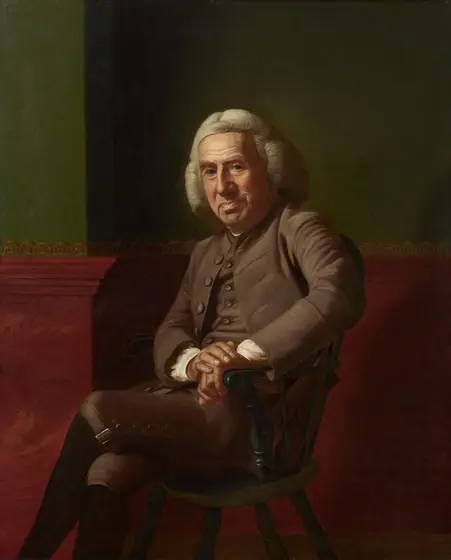 A pale-skinned, clean-shaven man with a heavily lined, paunchy face sits against a green and red wall in this vertical portrait painting. The man sits in a wooden chair painted forest green and rests both hands on the closer arm.