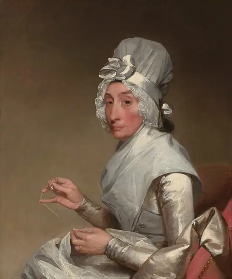 Shown from the lap up, a woman with pale skin wearing a white satin dress and tall white bonnet sits sewing with her body facing our left in this vertical portrait painting.