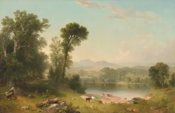 We look sightly down and across a green meadow in front of us at a grove of tall trees to our left next to a shimmering body of water, before a deep view of distant mountains beneath an ice-blue sky in this horizontal landscape.